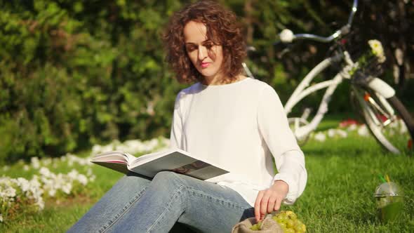 Young Woman in White Shirt and Blue Jeans is Reading a Book Sitting on the Grass in Park and Eating