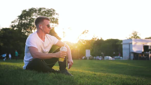 Handsome Young Hipster Man with Earphones Outdoors in Park with Lemonade in His Hand