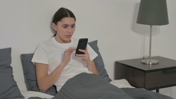 Hispanic Woman Reacting to Loss on Smartphone in Bed