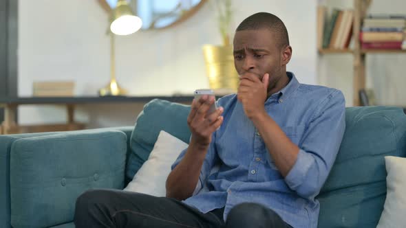 Young African Man Reacting To Loss on Smartphone on Sofa 