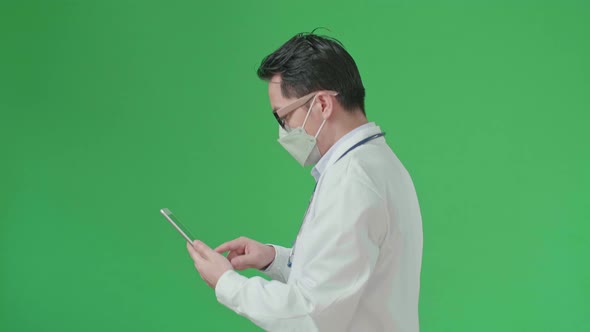 Asian Doctor With Stethoscope Wearing Mask, Using Tablet While Walking On Green Screen