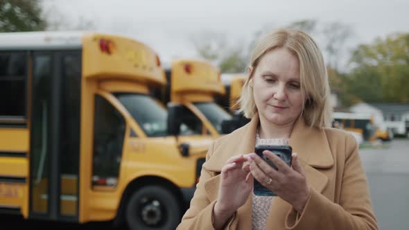 A Teacher Uses a Smartphone Stands in the School Yard Against the Background of Yellow School Buses