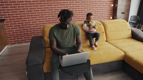 Busy Young African Father Doing Freelance Work on Laptop and Babysitting Young Child at Home