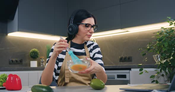 Woman in Earphones Enjoying Music while Whipping Eggs in Bowl Using whisk at the Table 