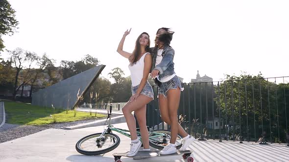 Two Cuddy Jovial Sexy Girls in Fashion Clothes Standing by One Leg on Skateboard with Raised Hands