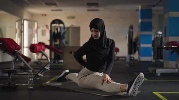 Wide Shot Serious Slim Fit Sportswoman in Hijab Stretching Leg Muscles Sitting on Twine Raising Leg