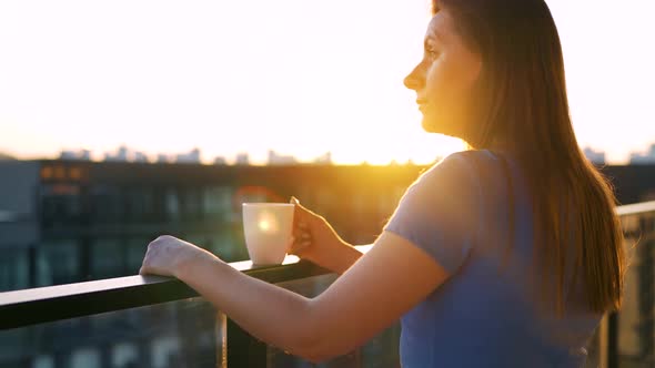 Woman with a Cup of Coffee Standing on the Balcony and Admire the Sunset