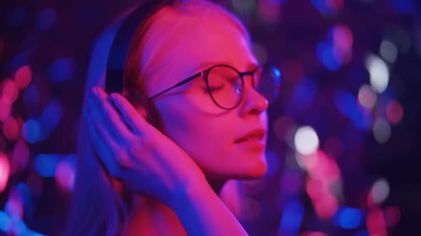 Happy Young Blonde Woman in Glasses Dances in Headphones on a Background of Multicolored Lights