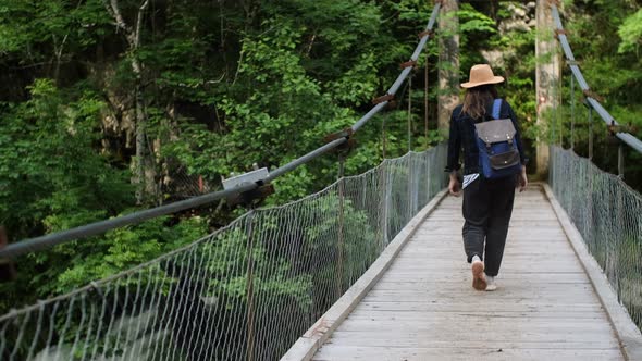 Back view of Hipster woman in hat with backpack walks on a suspended bridge over a mountain