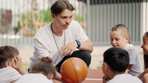 Sporty Coach Catching Ball and Explaining Game Strategy to Primary School Children in Basketball