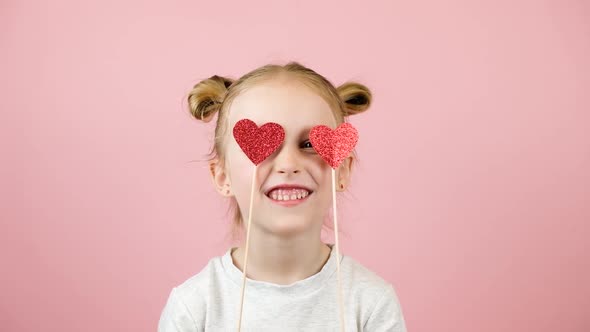 Funny Little Blonde Girl Smiling and Playing with Red Heart Toy on Pink Background