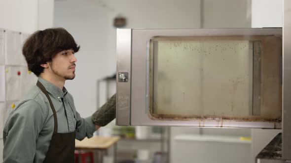 Young Man Baker in Apron and Glove Taking Fresh Pastry Out of Oven in Bakery Kitchen