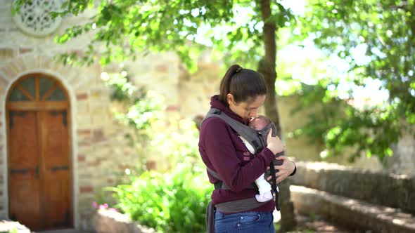 Mom Hugs and Kisses a Newborn Baby in a Sling in the Park