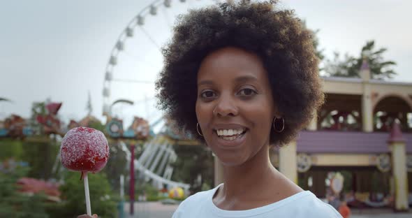 Portrait of Young African Woman Holding Candy Apple and Smiling at Camera in Amusement Park