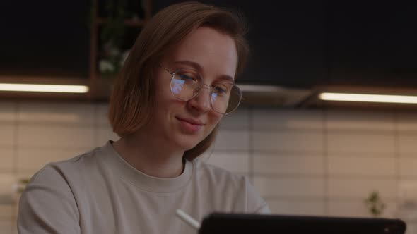 Smiling Girl in Glasses Studying at Home in the Kitchen Using a Tablet and a Digital Pen
