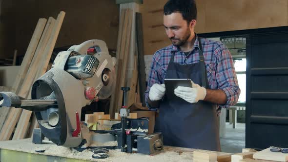 The Carpenter Works on Tablet Near Electric Saw in Workshop