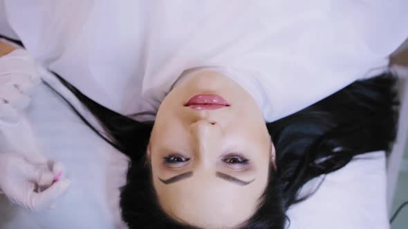 beautiful woman with black hair is lying on a couch on the procedure of permanent makeup lips