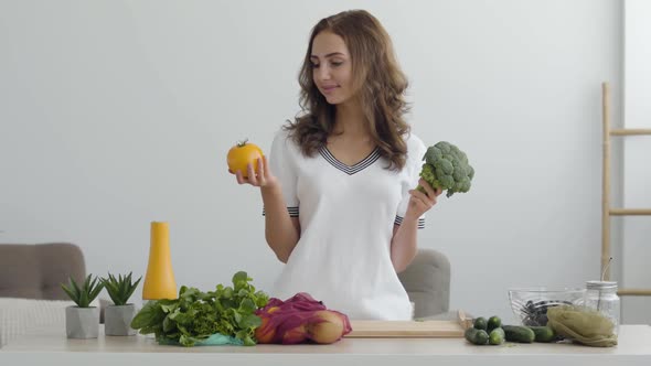 Young Smiling Woman Holding Yellow Tomato and Broccoli Standing at the Table in Modern Kitchen