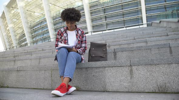 Pretty Afro-American Female Student Doing Homework Outdoors, Smiling Into Camera