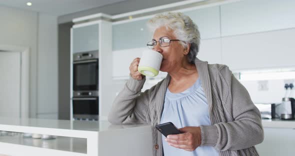African american senior woman drinking coffee and using smartphone in the kitchen at home