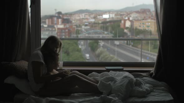 View of Mother Lying on the Bed with Small Son Against Huge Panoramic Window and Cityscape