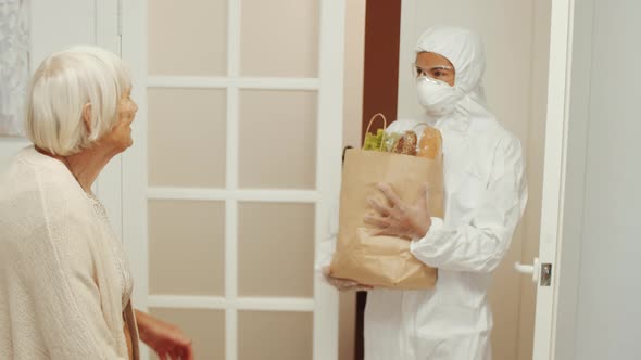 Caregiver in Protective Suit Bringing Groceries to Old Woman at Home