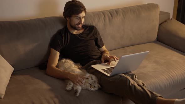 Happy Man Working on a Laptop at Home While Sitting on the Couch and Petting a Cat