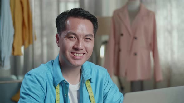 Asian Male Designer Working On A Laptop And Smiling To The Camera While Designing Clothes