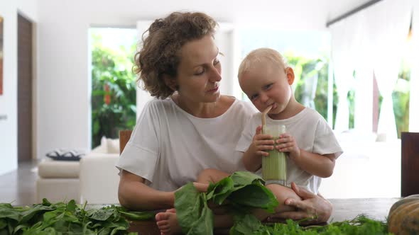 Caucasian Baby Sitting on the Kitchen Table Among Green Leaves Drinking Green Smoothie They've Just