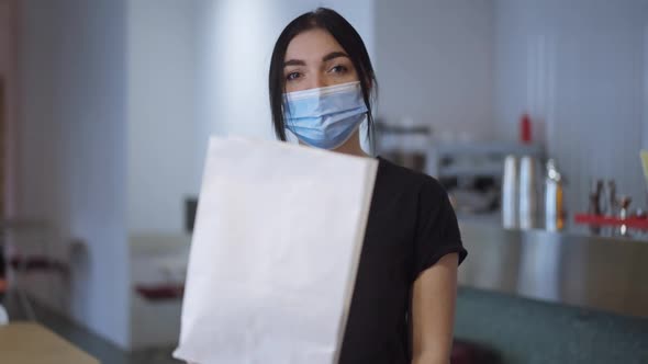 Medium Shot of Young Confident Woman in Covid Face Mask Stretching Packed Lunch to Camera Standing