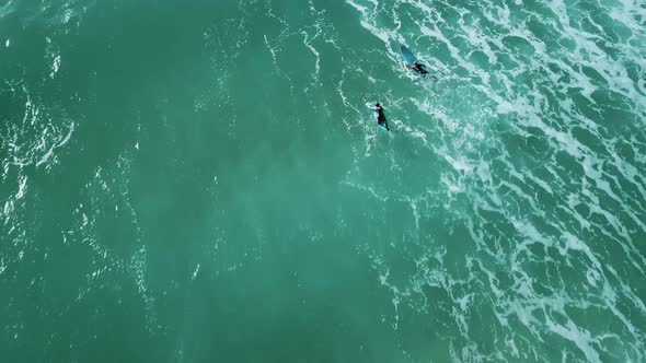 Drone shot of two surfers paddling together over waves.