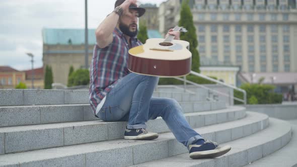 Caucasian Man Walking Up Urban Stairs, Taking Off Hat, and Start Playing Guitar. Portrait of Street
