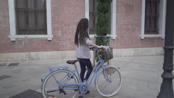 CurlyHaired Lady With Bicycle Is Walking Along The Sidestreet Of City Park