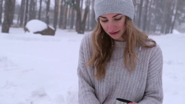 Cute Caucasian Girl in a White Sweater and Hat in a Snowy Forest Park Drinking Hot Tea or Chocolate