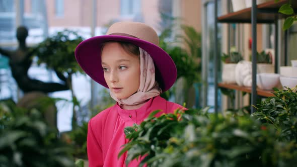 Young Cute Girl in Hat and Pink Jacket Looking Green Plants in Orangery