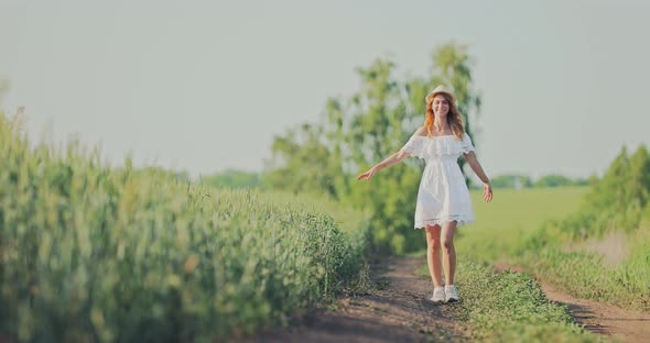 Beautiful Girl in a White Dress and Hat is Walking Along a Rural Road