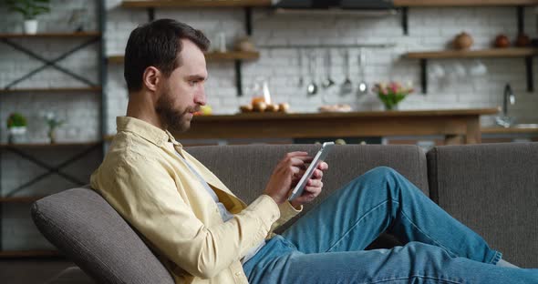 Young Man Using Digital Tablet Resting on Couch at Home Young Male Holding Computer Looking at