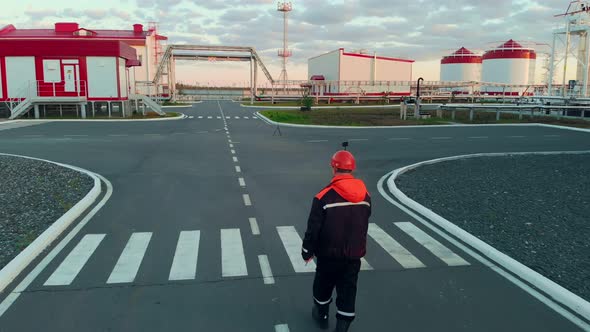 A Drone Follows a Technologist Wearing an Orange Hard Hat to an Oil and Gas Refinery