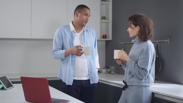 Young Married Couple Standing in Kitchen with Coffee Cups in the Morning Talking