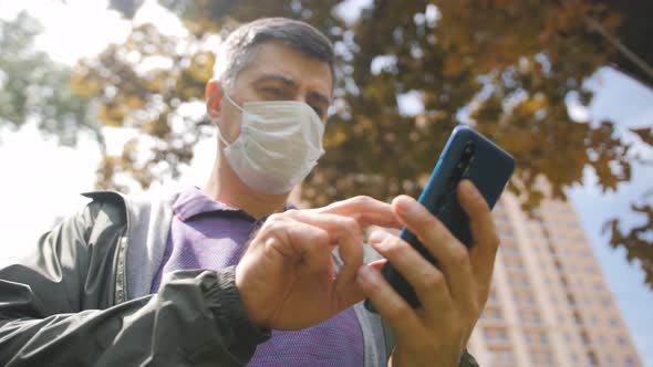 A Middleaged Man in a Medical Protective Mask is Talking on the Phone While Standing on a City