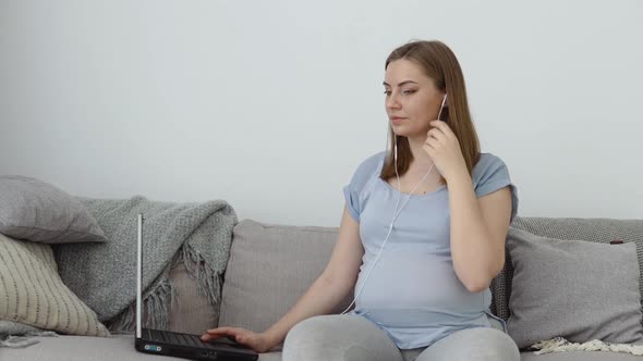 A Pregnant Woman in Home Clothes is Sitting on a Sofa and Using a Laptop