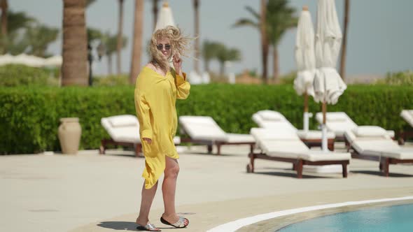 Sexy Blonde Lady in Yellow Dress is Walking Near Large Swimming Pool in Luxury Hotel