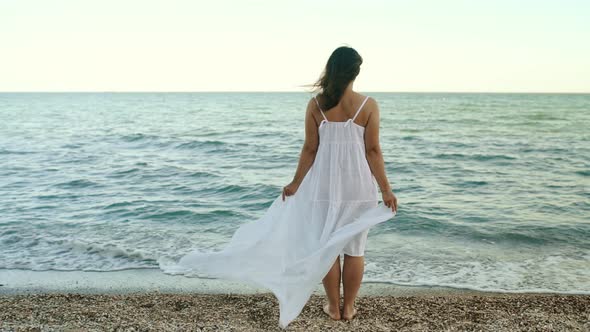 Young Woman in a White Dress Stands on the Seashore and Looks Into the Distance