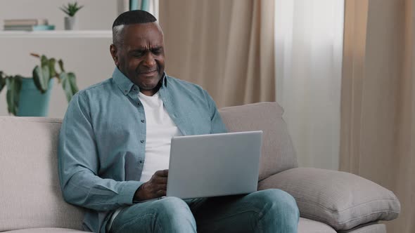 Mature Frustrated African American Man Sitting on Couch in Room Looks at Laptop Screen Reading Bad