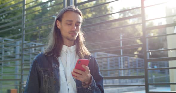 Long Haired Young Guy Chatting on Smartphone While Standing in Sunlight Near Sports Ground