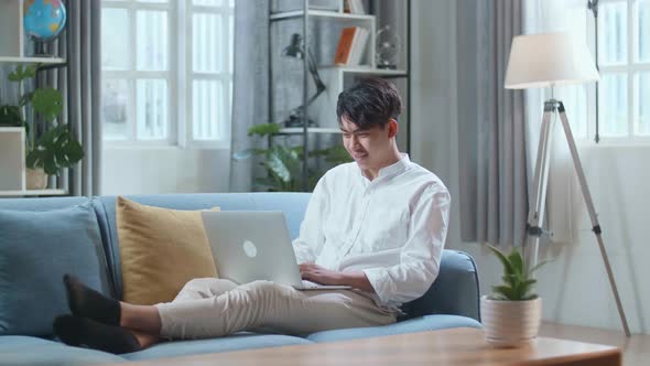 Full Body Of Smiling Asian Man Typing On Laptop Computer While Lying On Sofa In The Living Room