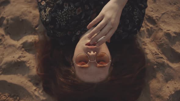 Sad Red-headed Girl Hipster Smokes a Cigarette While Lying on the Sand with His Eyes Closed. Pretty