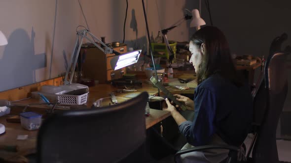 Focused caucasian female jeweller sitting at desk, making jewelry in workshop