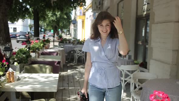 An Enthusiastic Young Woman Walks Along the Landing of a Summer Cafe Between Tables with a Camera in