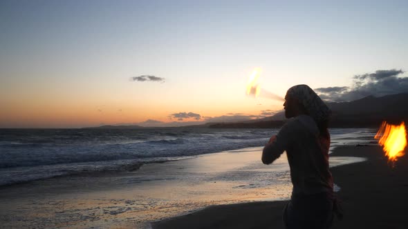 A man in silhouette dancing and spinning a burning fire staff on the beach at sunset with flames and